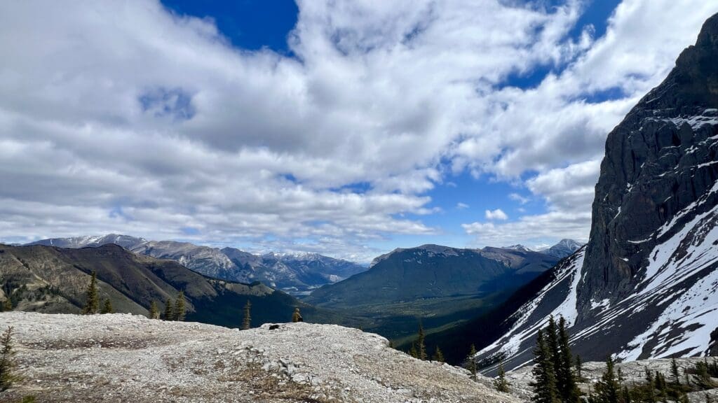 Expansive mountain landscape with rugged peaks, snow patches, and a deep valley. A hiker sits on a rocky ledge of West Wind Pass in Kananaskis, Alberta under a partly cloudy sky.