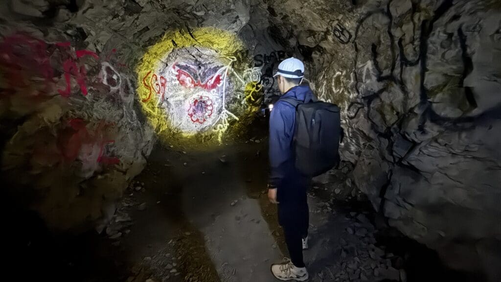 A person with a backpack and cap shines a flashlight on colorful graffiti on a dark cave wall inside Heart Creek Bunker in Kananaskis, Alberta. The scene feels mysterious and adventurous.