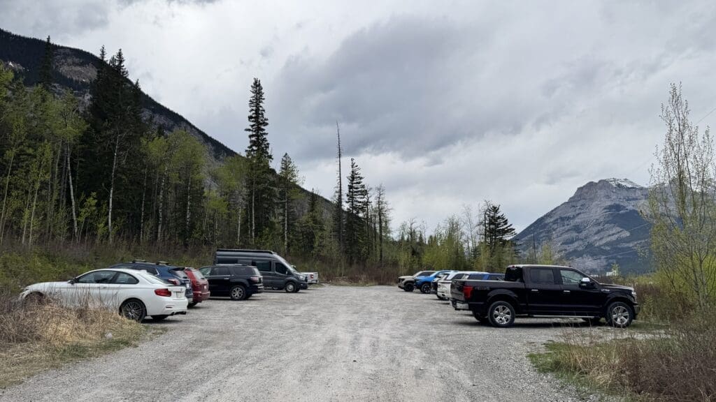 Gravel parking lot of Heart Creek Bunker Trailhead Parking in Canmore, Alberta with several parked cars and trucks. Surrounded by lush trees and mountains under a cloudy sky