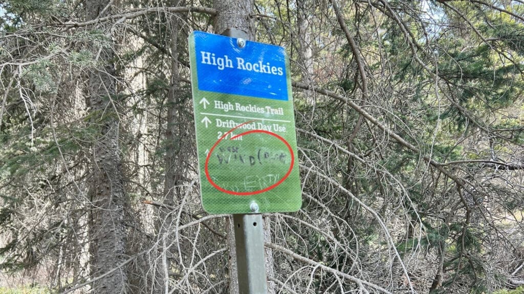 Signpost in a forest clearing reads "High Rockies." It includes directions to "High Rockies Trail", "Driftwood Day Use 2.7 km." and a hand-written direction to West Wind Pass in Kananaskis, Alberta. Surrounded by pine trees.