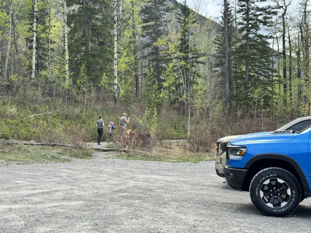 A blue truck is parked on a gravel area near a lush forest. Parents with kid and a dog walk toward the trees, starting to hike the Heart Creek Bunker Trail in Canmore, Alberta