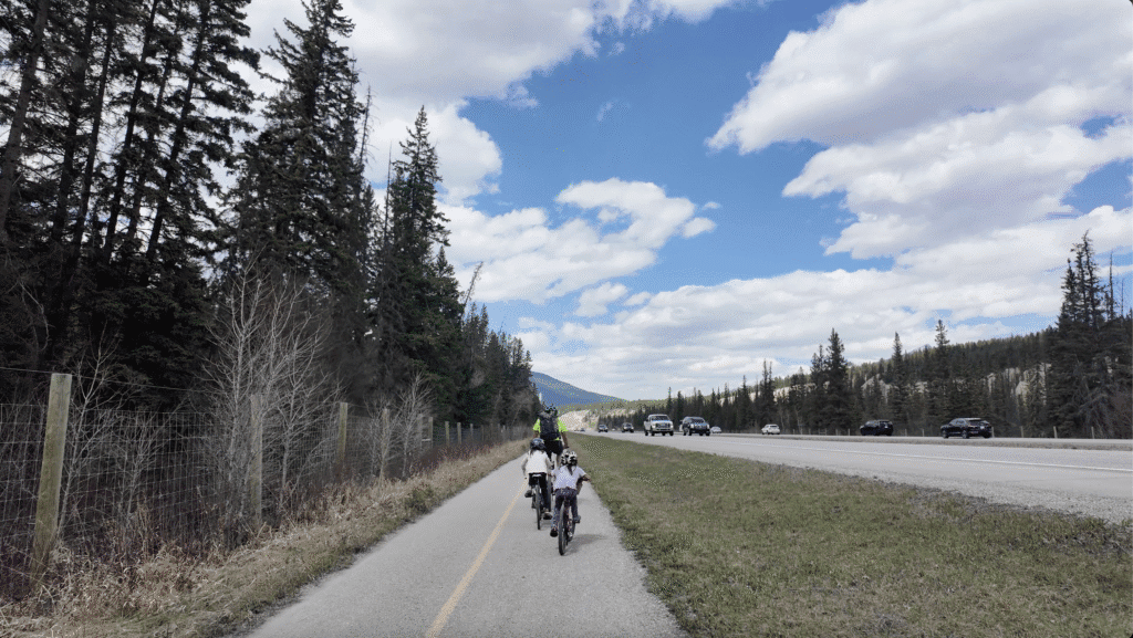 Two kids and their parent are biking on Legacy Trail in Banff National Park with tall trees on the left and a highway with cars on the right, under a partly cloudy blue sky.