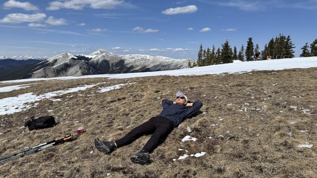 Hiker lies on dry grass near a snow patch on Prairie Mountain Summit in Kananaskis, with trekking poles and a backpack nearby, under a partly cloudy sky, with snowy peaks and trees in the background