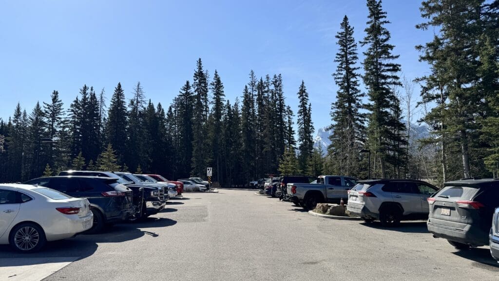 Parking lot at Legacy Trail in Canmore, Alberta with various cars on both sides, surrounded by tall pine trees under a clear blue sky
