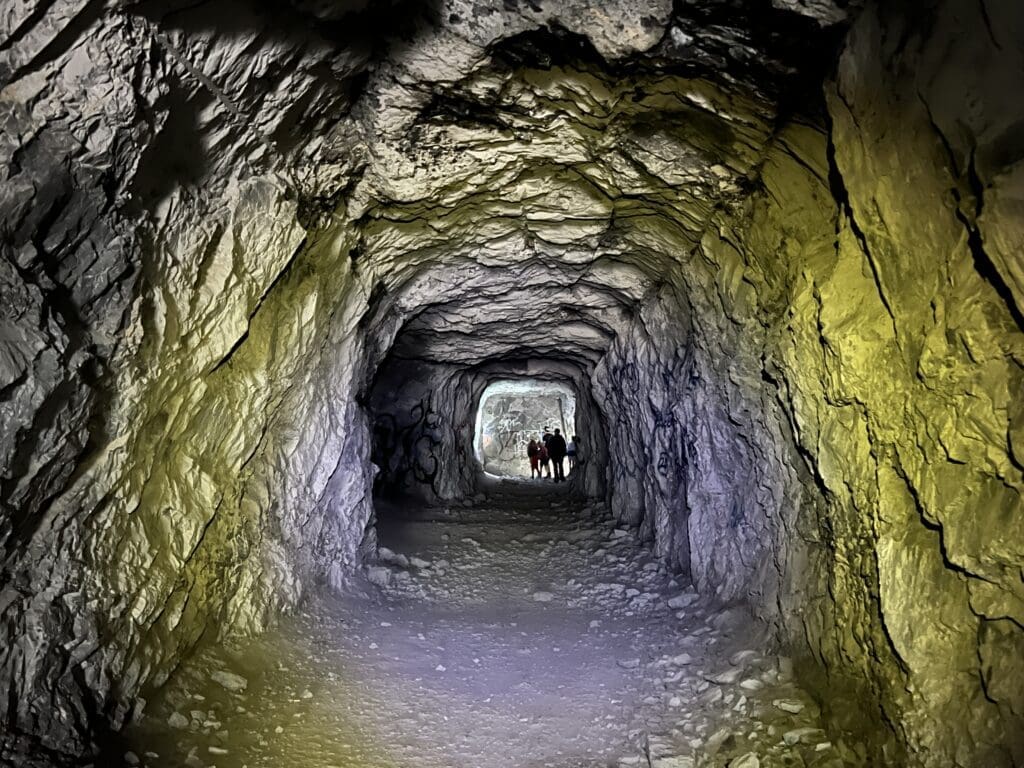 A dimly lit rocky tunnel of Heart Creek Bunker in Kananaskis, Alberta with rough walls in shades of yellow and gray. In the distance, a group of people walks towards the bright exit.