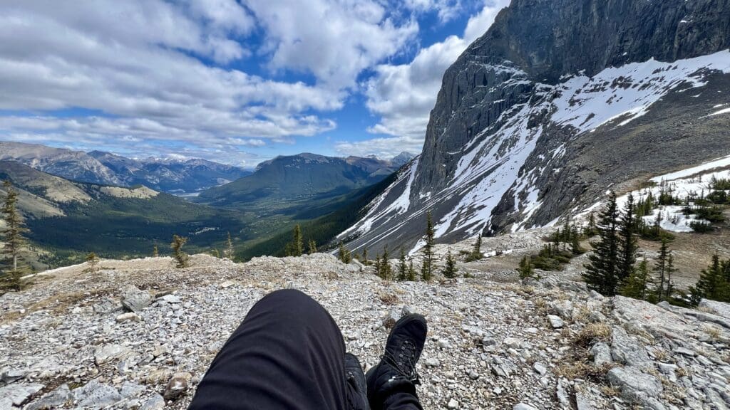View from a rocky ledge on West Wind Pass in Kananaskis, Alberta showing snow-capped mountains and a lush green valley. A person's legs in hiking boots are visible, evoking a sense of adventure.