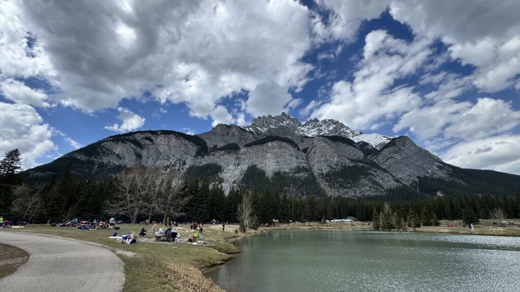 Mountain landscape with a scenic Cascade Ponds in the foreground. People enjoy picnics on grassy shores under a partly cloudy sky, with Cascade Mountain at the back in Banff National Park