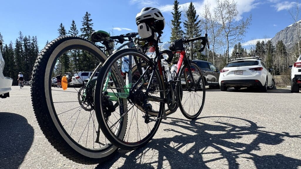 Bicycles parked in a sunny lot at Legacy Trail parking lot in Canmore, Alberta surrounded by cars and trees, with a white helmet rests on a handlebar, suggesting an outdoor cycling activity