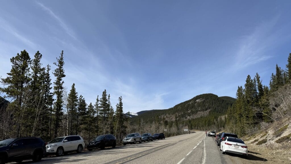 Vehicles parked along the roadside at Prairie Mountain trailhead parking area near Bragg Creek, Alberta, with forested foothills and distant mountain views under a clear blue sky.
