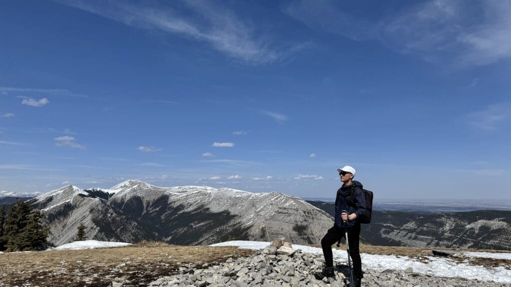 Hiker standing at the top of Prairie Mountain in Kananaskis with a pile of stones next to him, overlooking snow-capped ridges and a vast landscape under a clear blue sky