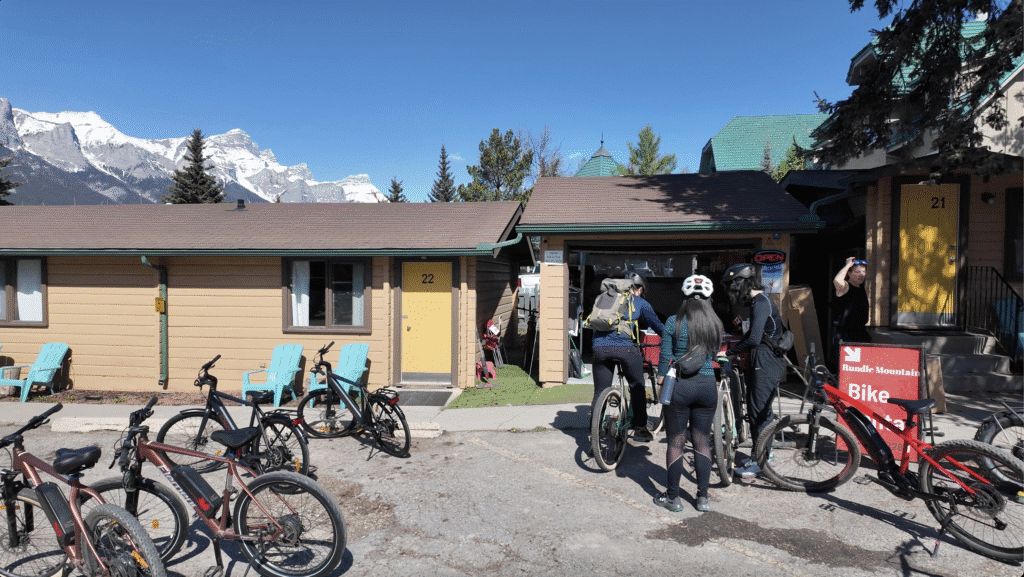Cyclists gather near a cabin of Rundle Rental Bike in Canmore with bright yellow doors, preparing for a ride. Snow-capped mountains loom in the background under a clear blue sky.