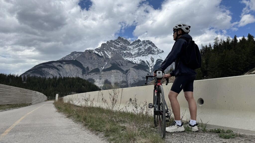 A biker wearing a helmet and backpack stands at Legacy Trail in Banff National Park beside a road bike, looking at the snow-capped Cascade Mountain under a partly cloudy sky, surrounded by dense forest