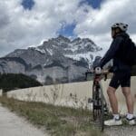 A biker wearing a helmet and backpack stands at Legacy Trail in Banff National Park beside a road bike, looking at the snow-capped Cascade Mountain under a partly cloudy sky, surrounded by dense forest