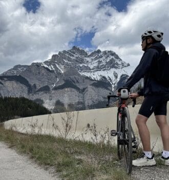 A biker wearing a helmet and backpack stands at Legacy Trail in Banff National Park beside a road bike, looking at the snow-capped Cascade Mountain under a partly cloudy sky, surrounded by dense forest