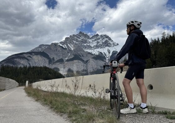 A biker wearing a helmet and backpack stands at Legacy Trail in Banff National Park beside a road bike, looking at the snow-capped Cascade Mountain under a partly cloudy sky, surrounded by dense forest