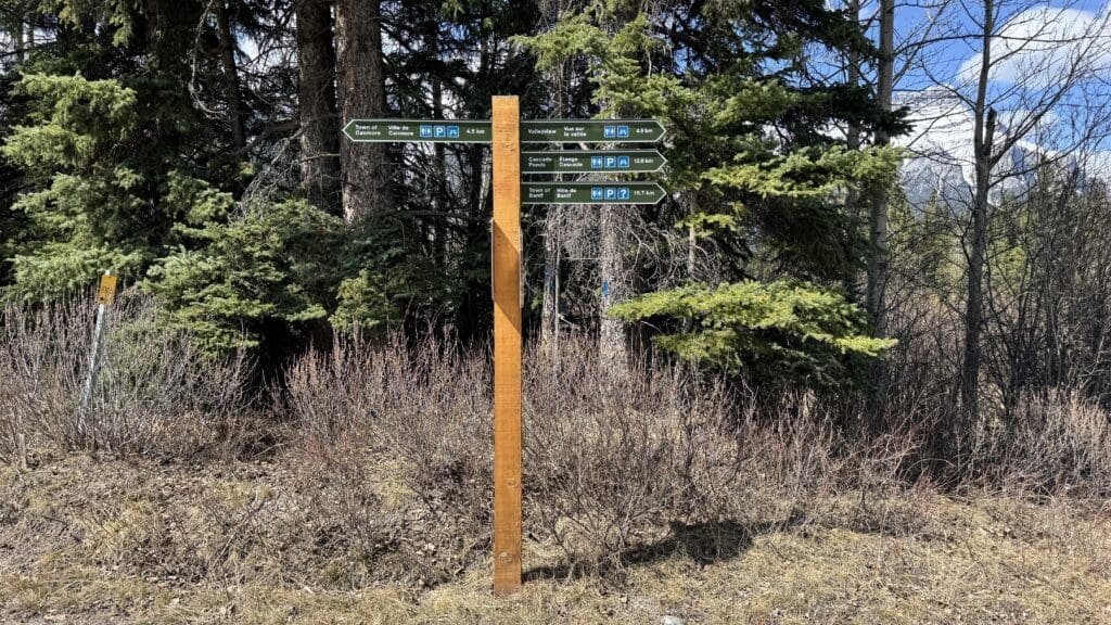 Wooden signpost with directional signs pointing to Canmore Town, Valleyview, Cascade Ponds, and Banff Town, set against a forest backdrop under a clear sky