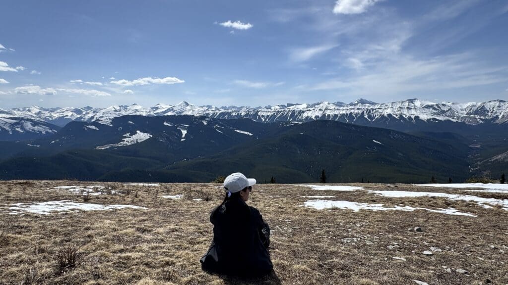 Hiker sitting at the summit of Prairie Mountain near Bragg Creek in Alberta Canada overlooking snow capped peaks and forested valleys of the Canadian Rockies under a clear blue sky