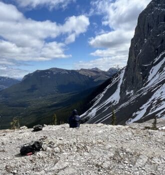 Person sits on rocky mountaintop of West Wind Pass in Kananaskis, Alberta, facing expansive mountain range under cloudy blue sky. Snow patches on steep cliff; serene, contemplative mood.