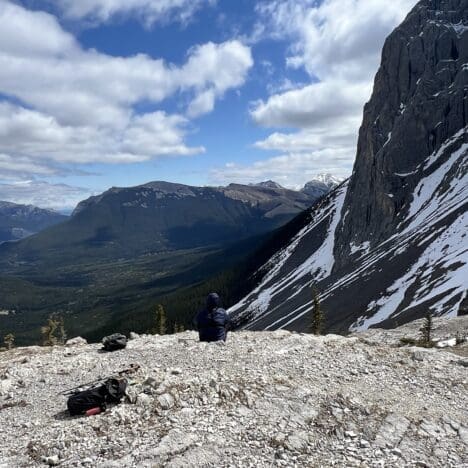 Person sits on rocky mountaintop of West Wind Pass in Kananaskis, Alberta, facing expansive mountain range under cloudy blue sky. Snow patches on steep cliff; serene, contemplative mood.