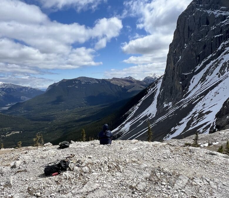 Person sits on rocky mountaintop of West Wind Pass in Kananaskis, Alberta, facing expansive mountain range under cloudy blue sky. Snow patches on steep cliff; serene, contemplative mood.