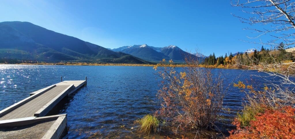 A serene Vermilion Lake in Banff National Park with a wooden dock on the left, surrounded by autumnal trees. Majestic mountains and a clear blue sky provide a peaceful backdrop.
