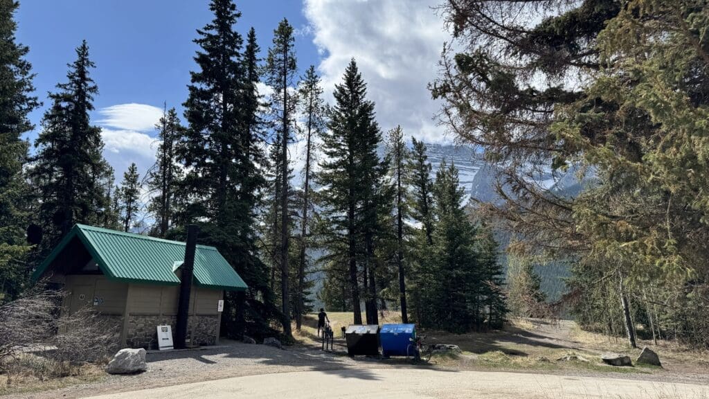 Washrooms with a green roof along Legacy Trail in Banff Natinal Park is surrounded by tall pine trees under a partly cloudy sky. A pathway leads into the forest, evoking a serene, natural setting.