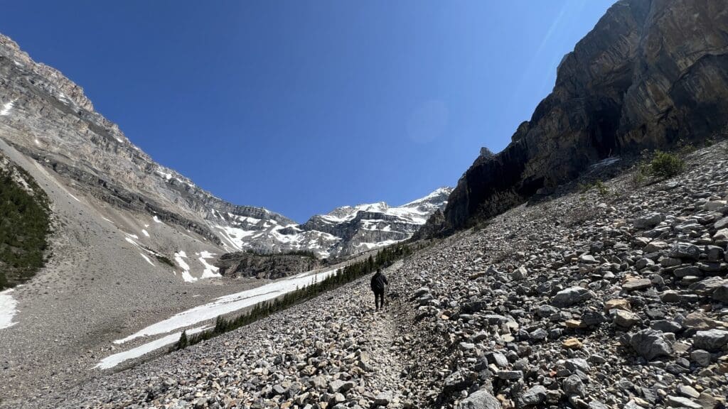 A lone hiker ascends the rocky mountain Stanley Glacier Trail in Kootenay National Park, BC under a clear blue sky. Snow patches cover parts of the rugged peaks, conveying a sense of serene adventure.