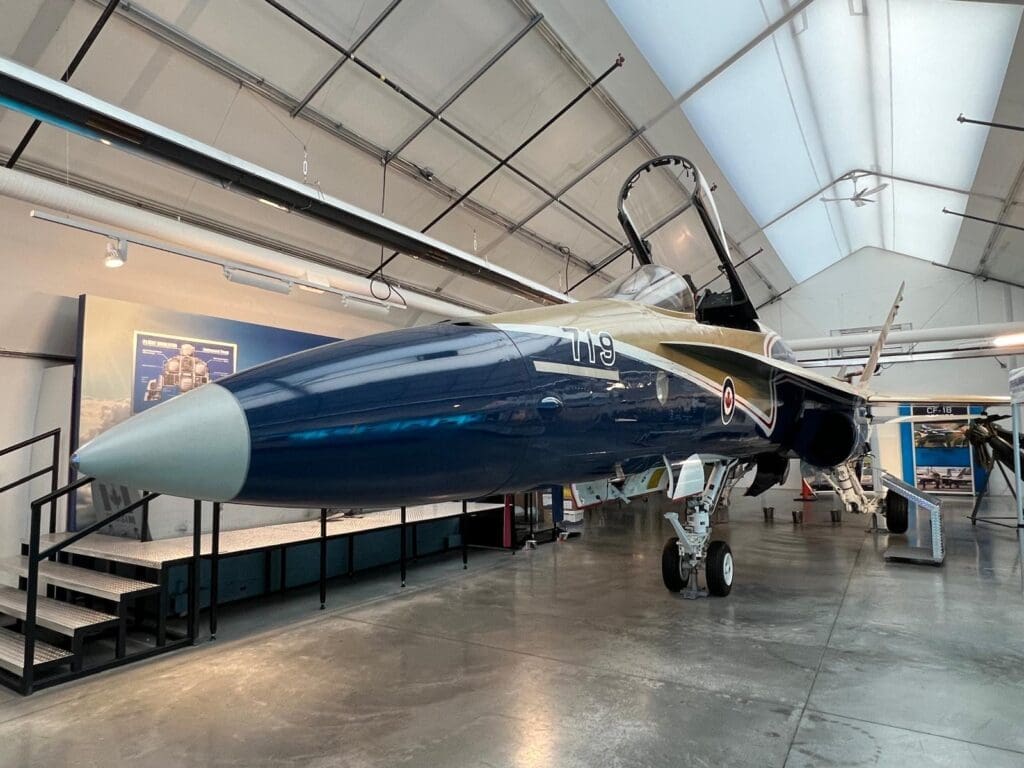 A blue and gold fighter jet displayed indoors in Military Museum in Calgary, Alberta, viewed from the side. The open cockpit and detailed landing gear create a dynamic and sleek appearance.