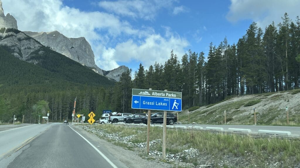 Road sign for Grassi Lakes in Kananaskis, Alberta in front of forested mountains under a blue sky with clouds. Cars are parked by the roadside, suggesting a popular hiking area.