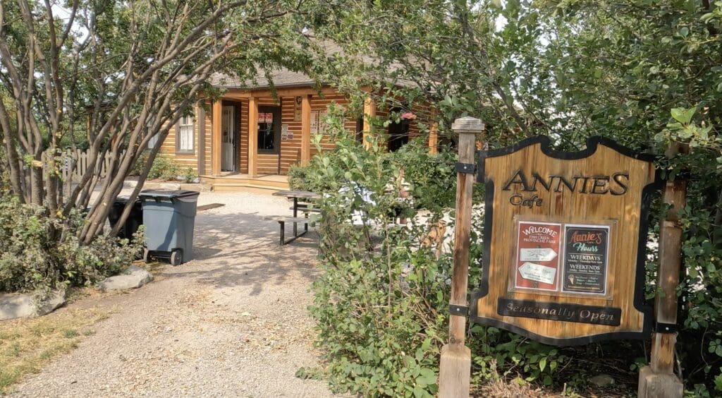 Charming log cabin-style café in Fish Creek Provincial Park in Calgary, Alberta partially obscured by leafy trees. A wooden sign reads 'ANNIE'S Cafe' and 'Seasonally Open.' Inviting atmosphere, rustic setting.