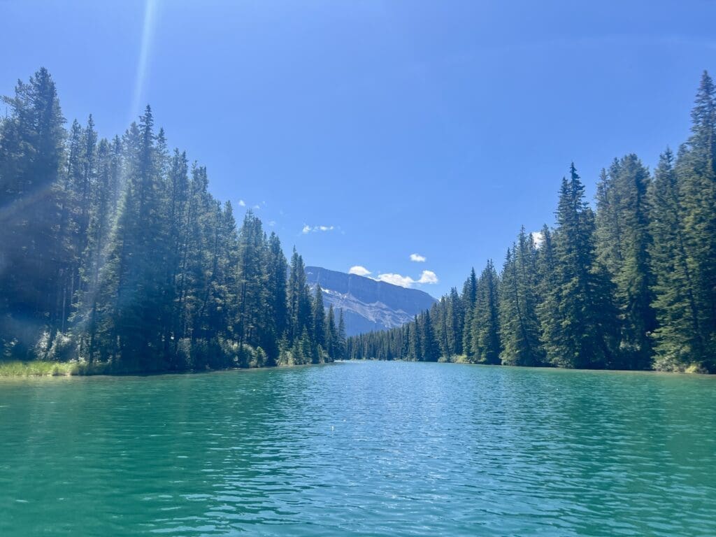 Two Jack Lake in Banff National Park, Alberta with clear turquoise water, bordered by dense green pine trees, leads to distant mountains under a bright blue sky with sunbeams.