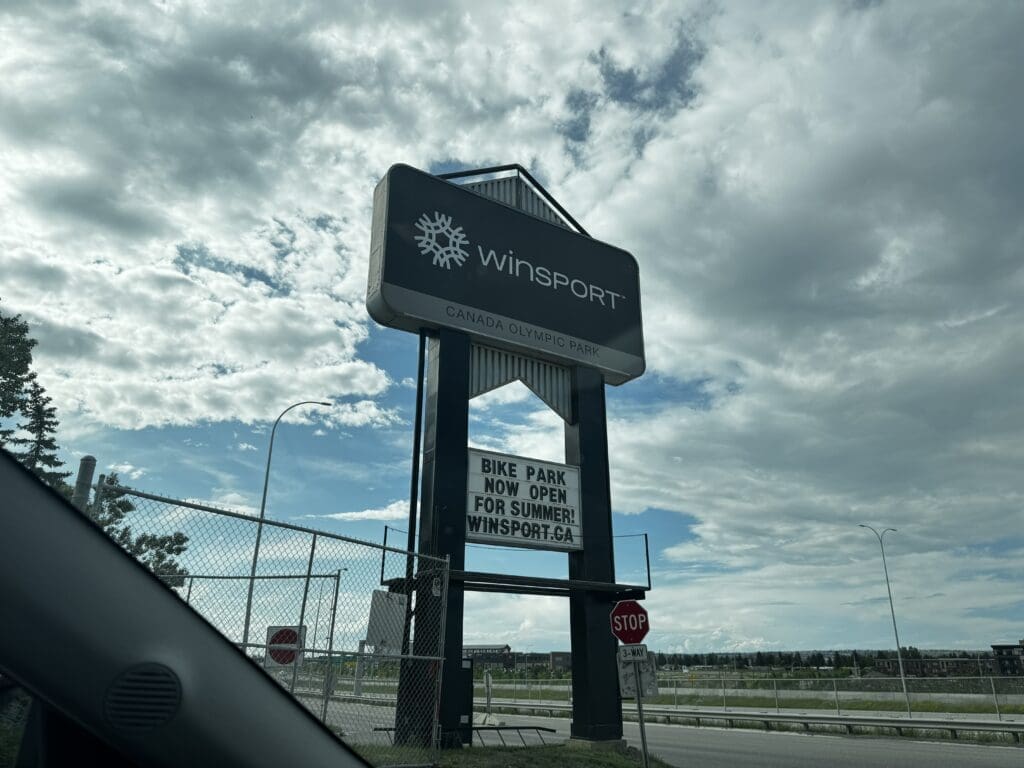 A large sign for WinSport Canada Olympic Park in Calgary, Alberta against a cloudy sky, promoting a summer bike park opening. A stop sign is visible in the foreground.