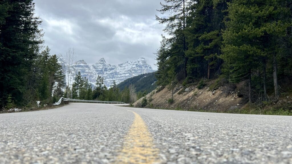 Mountain road perspective with a yellow centerline. Pine trees flank the Moraine Lake Road, while snow-capped peaks rise under a cloudy sky, creating a serene scene.