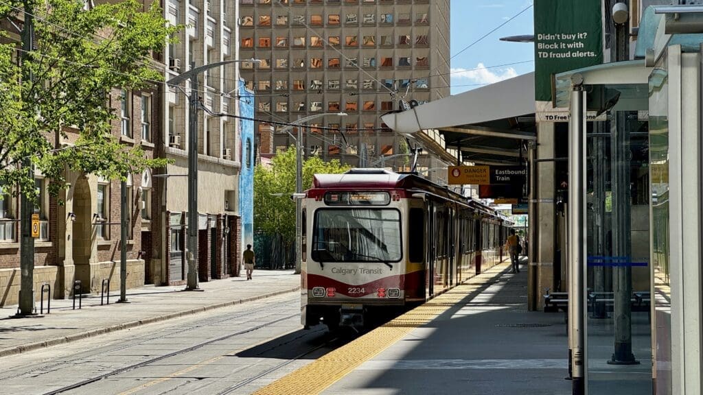 A Calgary Transit train in Alberta arrives at a sunny city street station. Tall buildings line the street, casting shadows. A person walks on the platform.