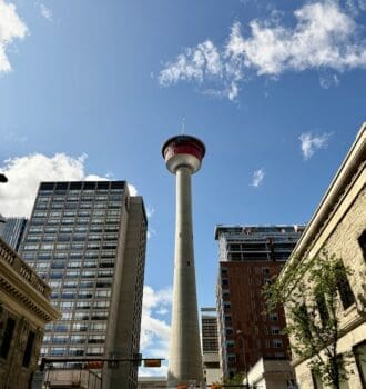 Tall Calgary Tower in Alberta under a blue sky with scattered clouds, surrounded by urban buildings. Cars and trees line the street, creating a vibrant cityscape.