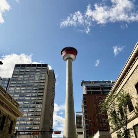 Tall Calgary Tower in Alberta under a blue sky with scattered clouds, surrounded by urban buildings. Cars and trees line the street, creating a vibrant cityscape.