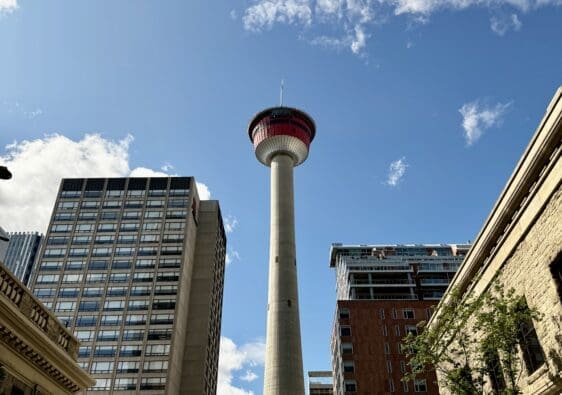 Tall Calgary Tower in Alberta under a blue sky with scattered clouds, surrounded by urban buildings. Cars and trees line the street, creating a vibrant cityscape.