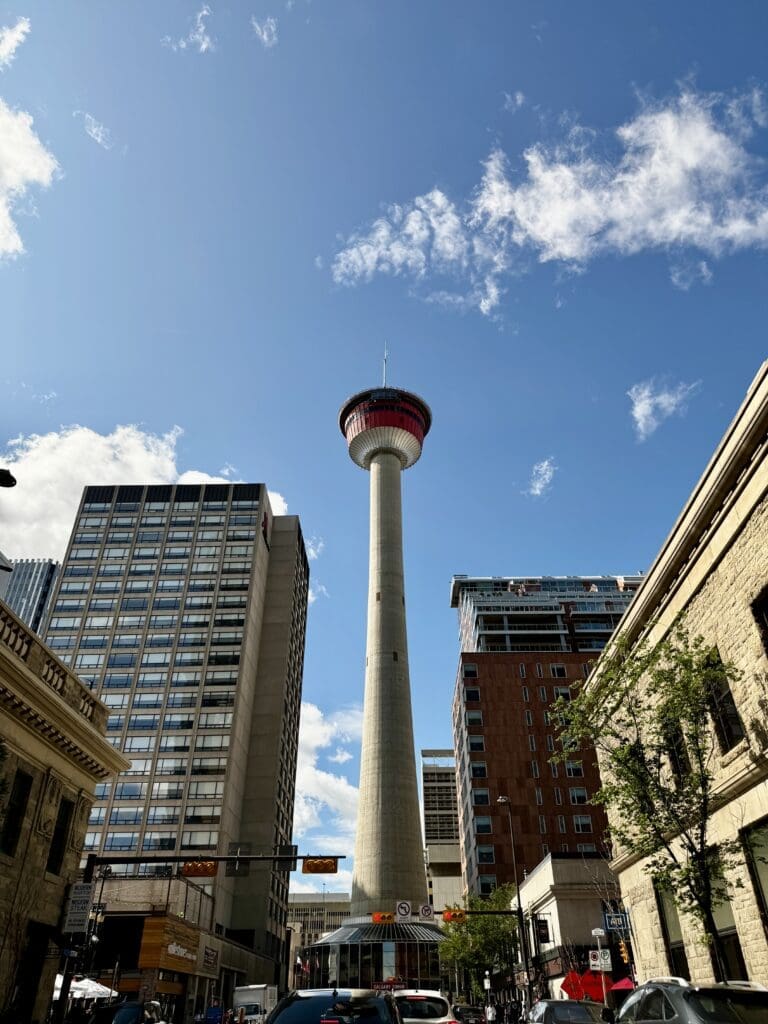 Tall Calgary Tower in Alberta under a blue sky with scattered clouds, surrounded by urban buildings. Cars and trees line the street, creating a vibrant cityscape.
