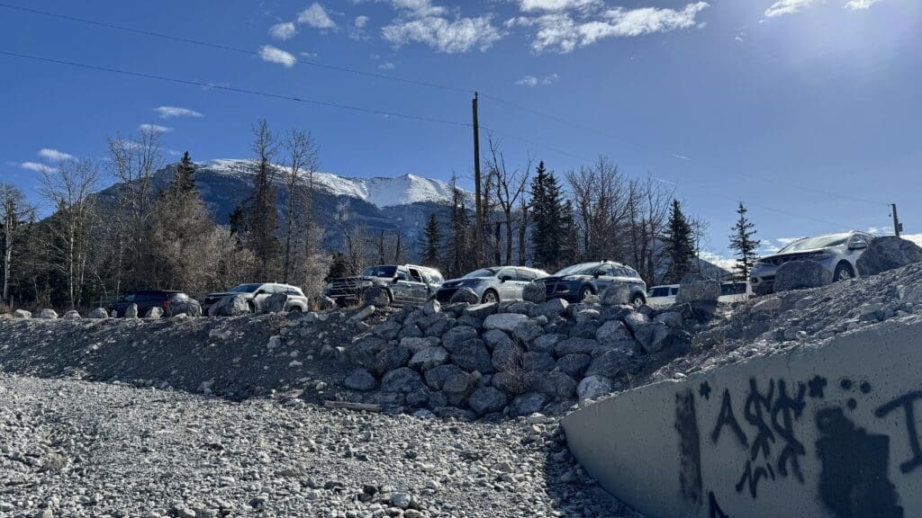Cars parked on a rocky hillside, with barren trees and a snow-capped mountain in the background under a clear blue sky. Foreground shows concrete graffiti.