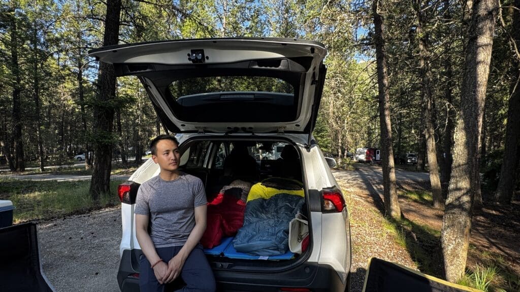 A man sits on the back of an SUV, open trunk filled with colorful sleeping bags, surrounded by a sunlit forest at Redstreak Campground in Kootenay National Park, BC.