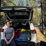 A man sits on the back of an SUV, open trunk filled with colorful sleeping bags, surrounded by a sunlit forest at Redstreak Campground in Kootenay National Park, BC.
