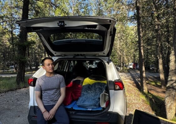 A man sits on the back of an SUV, open trunk filled with colorful sleeping bags, surrounded by a sunlit forest at Redstreak Campground in Kootenay National Park, BC.