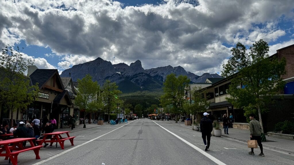 A lively street scene in the mountain town, Canmore, Alberta. People stroll and sit at red benches along the road. Majestic mountains under a cloudy sky in the background.