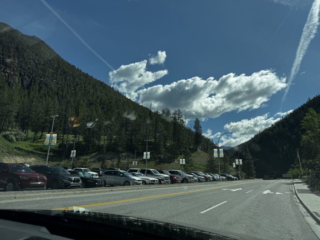 Roadside parking in Kootenay National Park, BC in front of a lush, green mountain under a bright blue sky with fluffy clouds. Several cars are visible, with banners on light poles.