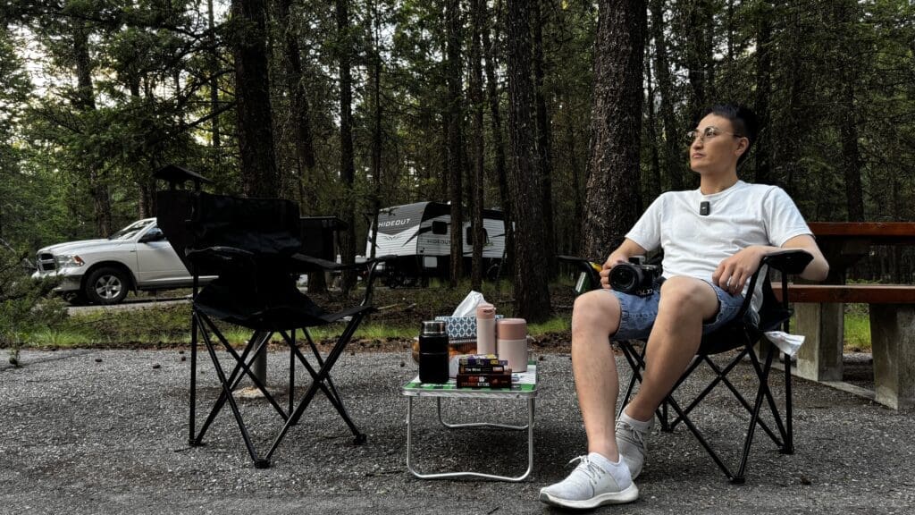 A person sits in a folding chair at Redstreak Campground in Kootenay National Park, BC, holding a camera, surrounded by trees. A white car and camper are in the background, creating a peaceful outdoor setting.
