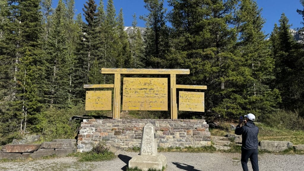 A person in a white cap photographs a large yellow sign amidst lush green pine trees. The sign marks the Continental Divide between Banff, Alberta and Kootenay National Parks, BC.