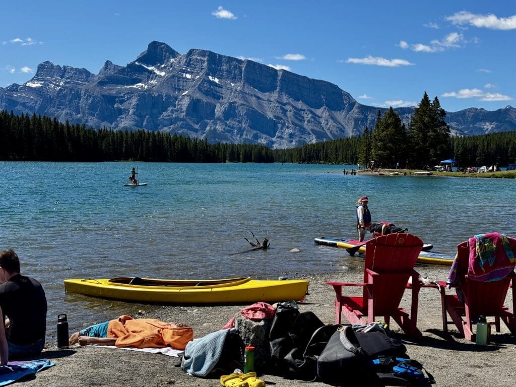 A serene lakeside scene with Two Jack Lake and majestic Mount Rundle in Banff National Park, Alberta in the background. People relax on the shore with kayaks and paddleboards nearby. Two red chairs add a splash of color, evoking a peaceful, leisurely atmosphere.