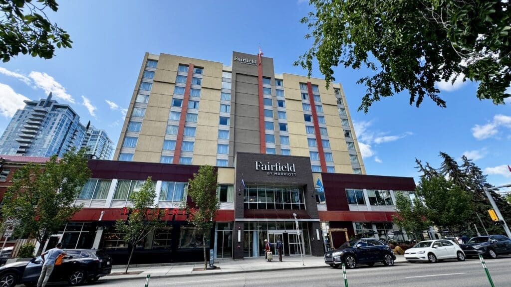 A mid-rise hotel building with "Fairfield by Marriott" signage in Calgary, Alberta on a sunny day, surrounded by trees and parked cars, reflecting an urban atmosphere.