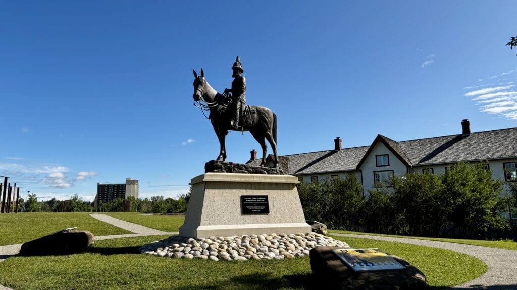 A bronze equestrian statue in Fort Calgary, Alberta on a pedestal is set against a bright blue sky. Nearby, a historic white house with a sloped roof. The scene exudes a serene and respectful atmosphere.