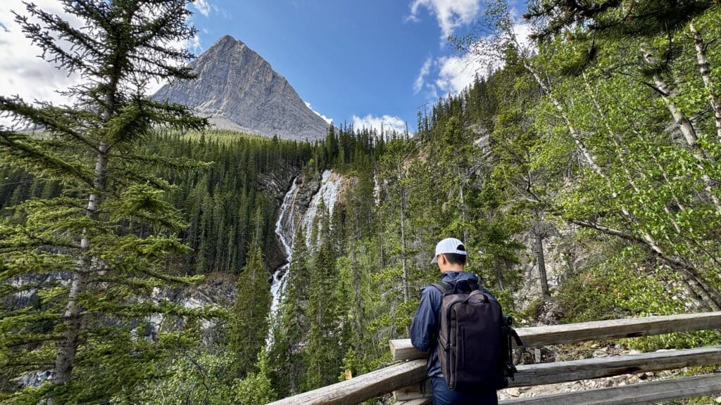 A person with a backpack and white cap stands on a wooden railing, overlooking a lush green forest and distant Grassi Falls in Kananaskis, Alberta under a blue sky with a rocky mountain.